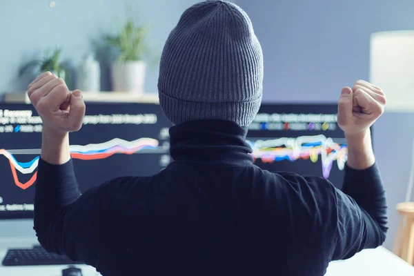 Back view portrait of hipster man broker looking at computer monitors and do winner gesture, clenched fists, celebrating successful trade on stock market, wearing gray cap.