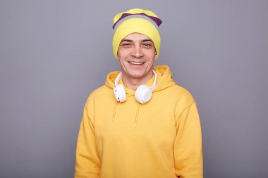 Portrait of attractive cheerful joyful man wearing yellow hoodie, beanie hat and headphones, looking at camera with pleasant smile, has friendly expression, standing isolated over gray background.