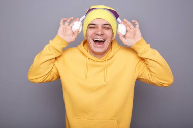 Image of satisfied smiling happy man wearing yellow hoodie and beanie hat holding his headphones, listening to music, laughing, standing isolated over gray background.