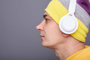 Profile portrait of serious concentrated man wearing yellow hoodie, beanie hat headphones standing isolated over gray background, looking aside with assertive face, copy space, advertisement area.