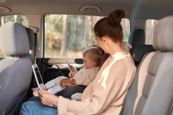 stock image Image of Caucasian woman working on laptop while sitting with her baby daughter in safety chair on backseat of car, child watching cartoons on tablet, family traveling.