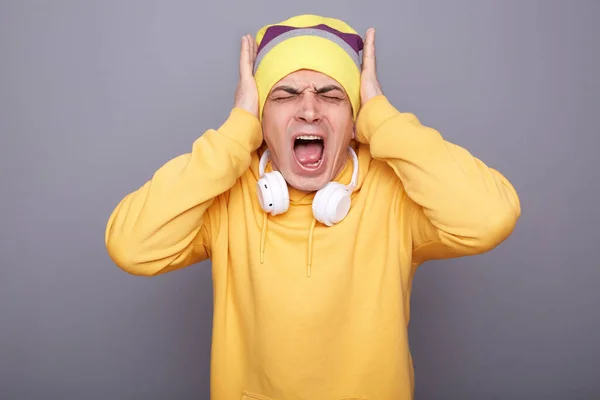 Portrait of shocked annoyed man wearing yellow hoodie, beanie hat and headphones, covering ears with hands, screaming, hearing loud noise, standing isolated over gray background.