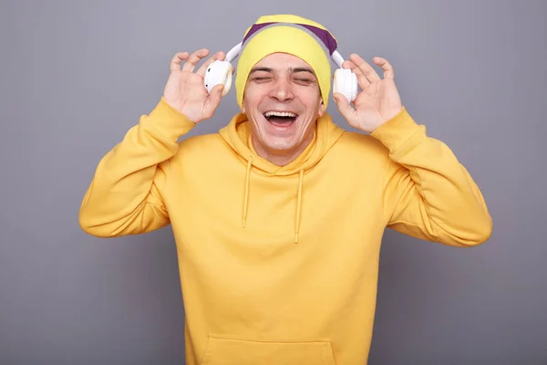 满意的笑脸 快乐的男人戴着黄色的帽帽 头戴耳机 听着音乐 在灰色的背景下孤立无援 — 图库照片