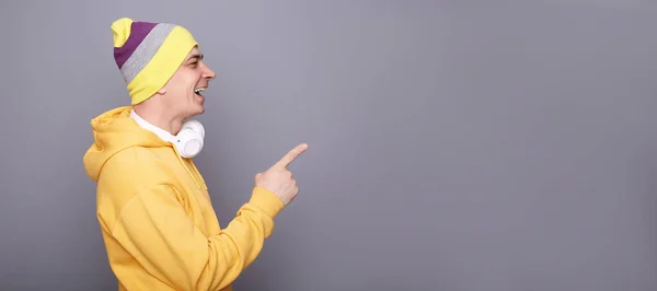 Side view portrait of laughing handsome man wearing yellow hoodie, beanie hat headphones standing isolated over gray background, pointing aside at copy space for advertisement or promotion.