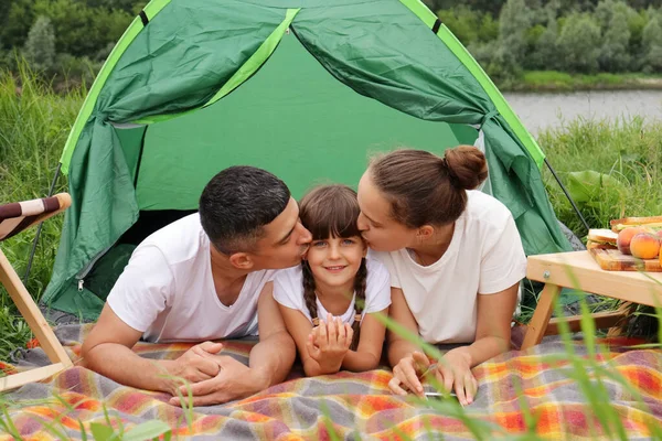 Image of loving family laying near the tent on blanket, mother and father kissing their daughter on cheeks, expressing positive emotions, having camping near the river in summer.