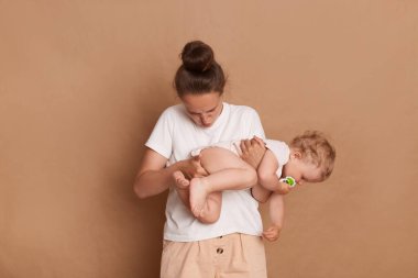 Photo of mom looking in stinky diaper of her little daughter, holding infant kid and does a grimace for disgust, standing isolated over brown background.