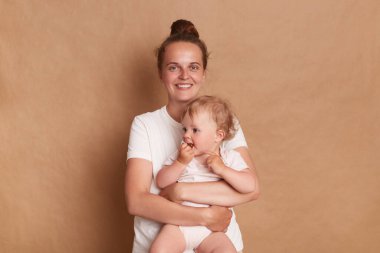 Portrait of delighted smiling woman with bun hairstyle wearing white T- shirt standing with her baby daughter isolated over brown background, looking at camera with happiness.