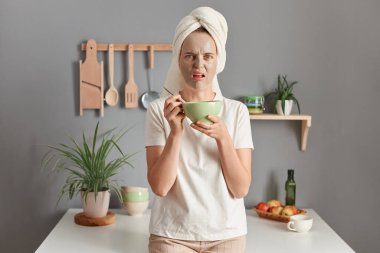 Photo of scared Caucasian woman with towel on head standing speechless indoor with beauty facial mask to reduce fine lines eats her breakfast, awakes early in morning, posing in kitchen.