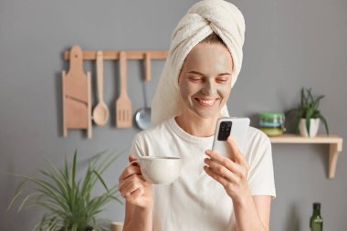 Indoor shot of smiling cheerful attractive woman wrapped in towel wearing white T-shirt standing in kitchen, using mobile phone, reading message or checking social network.