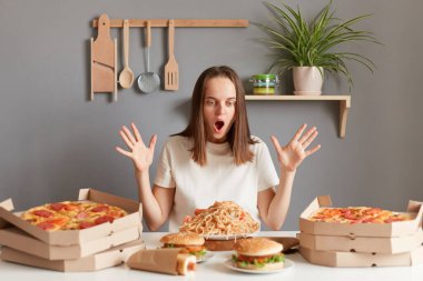 Photo of amazed excited woman with brown hair wearing white T-shirt sitting at table with raised arms and open mouth, being ready to eat pasta, hamburger, pizza and other junk food.