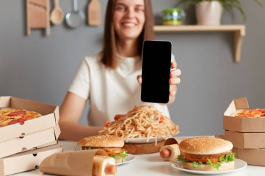 Kapalı alanda, kahverengi saçlı, beyaz tişörtlü, pizza, hamburger, makarna, hamur içinde sosis ve reklam için boş bir cep telefonuyla masada oturan oldukça olumlu bir kadının fotoğrafı..