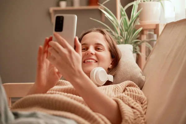 ソファに横たわっている間 スマートフォンを使用してヘッドフォンを身に着けている若い大人の魅力的な女性の屋内ショットは 喜んで笑顔 週末に無料の時間を楽しんで インターネットを閲覧 — ストック写真