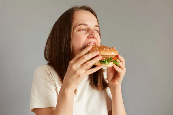 Profile portrait of Caucasian brown haired woman biting of appetizing delicious hamburger, being extremely hungry, enjoying junk food, finishing diet, having cheat meal.