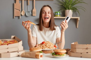 Indoor shot of amazed excited young adult woman with brown hair wearing white t shirt sitting in kitchen at table and looking at camera, holding in hands, smart phone and slice of pizza.