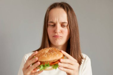 Portrait of dissatisfied sad woman wearing white T-shirt holding burger isolated on gray background, thinking eat or not, proper nutrition, healthy fast food, unhealthy choice.