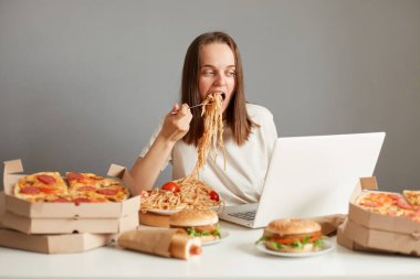 Kapalı alanda aç bir kadının laptopta çalışırken fast food yerken, gri duvara karşı masada otururken, dizüstü bilgisayarda çalışırken ve makarna yerken, ağzını açık tutarken, dizüstü bilgisayar ekranına bakarken..