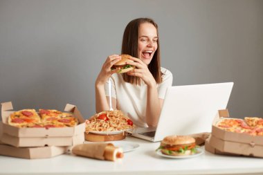 Tatmin olmuş Kafkasyalı kadın fast food yiyor, gri duvarda bir sürü abur cuburla masada oturuyor, elinde bir dilim pizza ve hamburger tutuyor, gözlerini kapalı tutuyor..