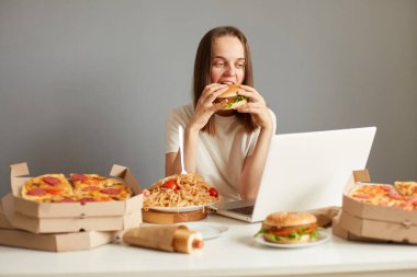 Koyu renk saçlı çekici bir kadının portresi fast food yiyor, gri duvara yaslanıp film seyrediyor, büyük hamburger yiyor, abur cubur yiyor..