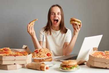 Dizüstü bilgisayardan film izlerken fast food yiyen heyecanlı genç bir kadının portresi gri duvara yaslanmış, elinde sandviç ve pizza dilimleri tutarak bağırıyor..