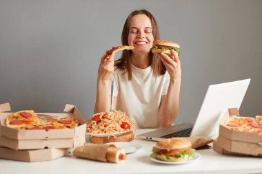 Tatmin olmuş Kafkasyalı kadın fast food yiyor, gri duvarda bir sürü abur cuburla masada oturuyor, elinde bir dilim pizza ve el burgeri tutuyor, gözlerini kapalı tutuyor..