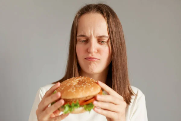 不満悲しい女性の肖像白いTシャツを着て灰色の背景に隔離されたハンバーガーを保持し 考えて食べるかどうか 適切な栄養 健康的なファーストフード 不健康な選択 — ストック写真