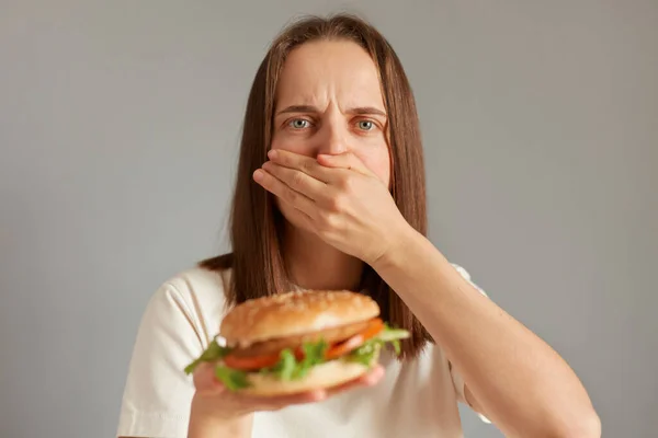 stock image Indoor shot of ill woman feeling sick when see fast food, covering her mouth feels nausea, eating spoiled sandwich, looking at camera, feeling unwell.