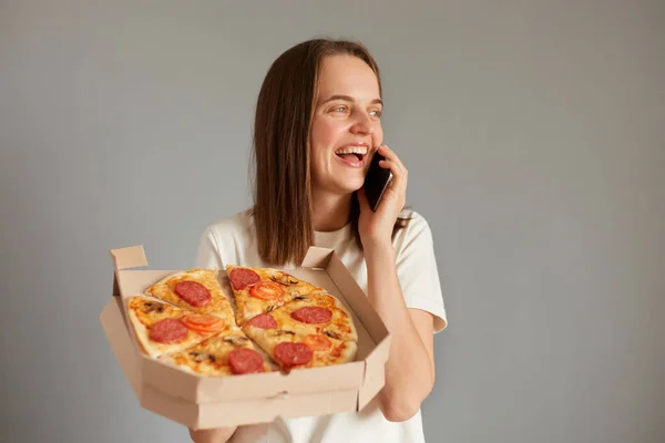 Indoor shot of laughing woman dressed white casual T-shirt standing holding take away pizza isolated over gray background, talking on mobile phone, inviting friend for having snack.