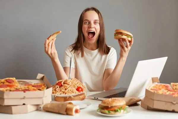 興奮した若い大人の女性の肖像画は ラップトップ上で映画を見ながら ファーストフードを食べて 灰色の壁に対してテーブルに座って ピザのサンドイッチとスライスを保持し 叫んで — ストック写真