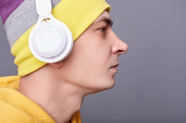 Yakından bakıldığında, bereli şapka takan ciddi bir adamın portresi ve gri arka planda izole edilmiş sarı bir kapüşonlu. Katı bir yüz ile ileriye bakıyor, kulaklıkla podcast dinliyor..