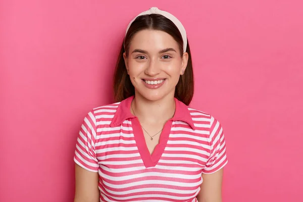Indoor shot of cheerful charming young girl with hair band wearing striped casual t shirt, looking at camera with toothy smile, standing isolated pink background, expressing positive emotions