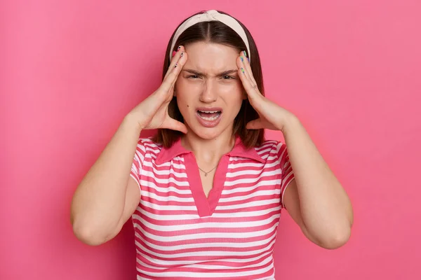 Indoor shot of irritated angry aggressive young girl with hair band wearing striped casual t shirt standing isolated pink background, screaming with angry, having headache