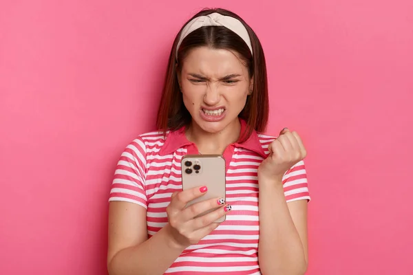 Horizontal shot of sad irritated young girl with hair band wearing striped casual t shirt, holding with smart phone in hands, reading crazy message, expressing anger, standing isolated pink background