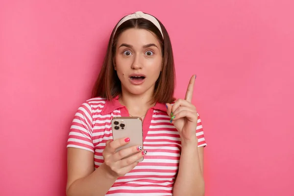 Photo of excited inspired shocked young girl with hair band wearing striped casual t shirt standing isolated pink background, using mobile phone, having idea about new app, raised finger up