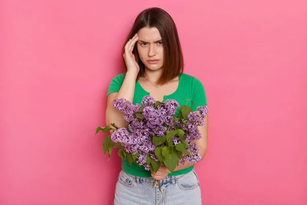 stock image Portrait of sick young girl with brown hair wearing green casual t shirt holding bouquet of lilac flowers, suffering headache, posing isolated over pink wall background