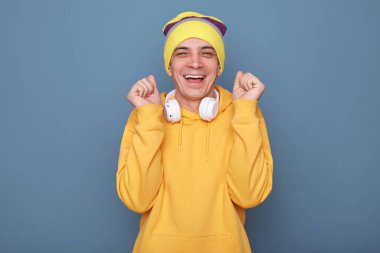 Son derece mutlu, bereli ve sarı kapüşonlu bir adam portresi, yumruğunu sıktı, başarısını kutladı, mutlu bir şekilde gülümsedi, mavi duvara karşı poz verdi.