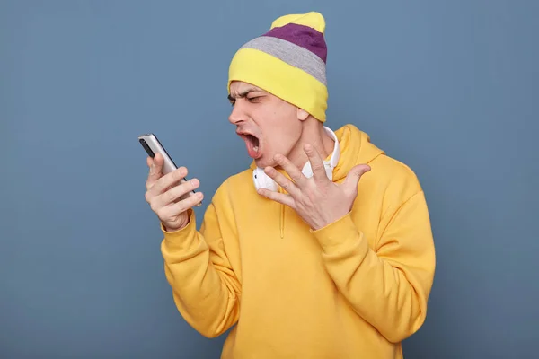 一个有侵略性的白人男子头戴便帽 头戴黄帽 用手机 阅读坏消息 怒气冲冲地尖叫 被蓝色背景隔离的形象 — 图库照片