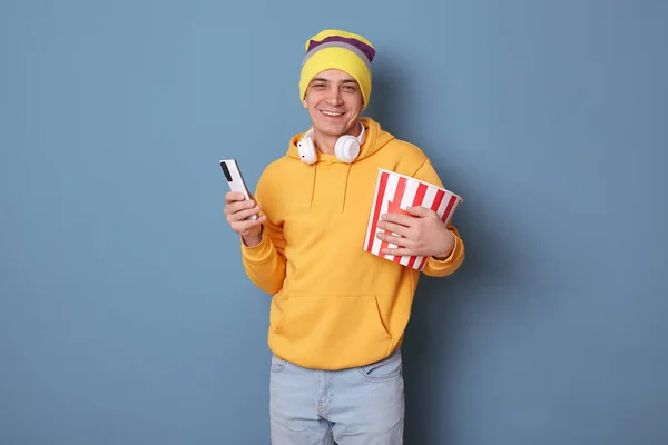 在室内拍摄的快乐英俊的高加索人 头戴便帽 头戴黄色休闲鞋 与蓝色背景隔离 看电影 拿着装有爆米花和智能手机的桶 — 图库照片