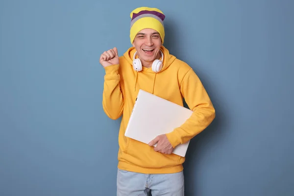 照片上的人喜形于色 笑容满面 头戴便帽 头戴黄帽 手持紧闭的笔记本电脑 握紧拳头 完成了一个项目 庆祝着 靠在蓝色的墙上摆姿势 — 图库照片