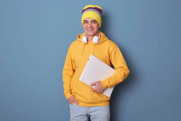 Indoor shot of delighted attractive man wearing beanie hat and casual yellow hoodie posing against blue wall, being very glad, waiting for starting work, expressing positive emotions