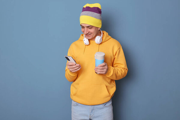 Крытый снимок улыбающегося блогера в шапочке-шапочке и непринужденной желтой толстовке, позирующей у голубой стены, пьющего горячий напиток из термоса и пользующегося смартфоном, общающегося с друзьями в социальных сетях