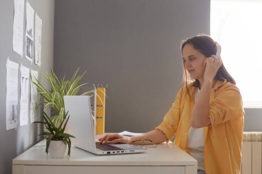 Çekici kahverengi saçlı bir kadının portresi. Masada oturuyor, dizüstü bilgisayarda çalışıyor ve kulaklıkla bir şeyler dinliyor, ekrana bakıyor, sarı gömlek giyiyor, hoş bir ifade takınıyor..