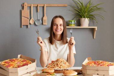 Kahverengi saçlı, beyaz tişörtlü, mutfaktaki masada oturan, çatalları elinde tutan, lezzetli makarna ve diğer fast food 'ları yemeye hazır gülümseyen yetişkin bir kadın resmi..