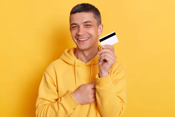 一个英俊的 面带微笑的黑发男子的画像 他在黄色背景下出示信用卡 面带微笑地看着相机 在购物中心用卡片付款 — 图库照片