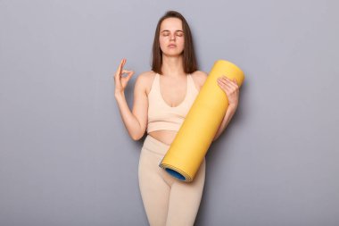 Bej bluzlu ve tozluklu sakin bir kadının gri arka planda izole edilmiş spor minderini tutarken, meditasyon yaparken, kapalı gözlerle yoga yaparken.