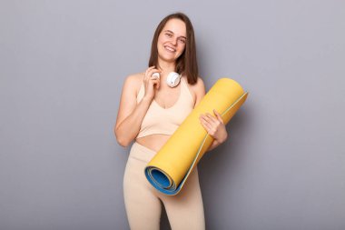Dost canlısı, keyifli bir kadının spor kıyafetleri içinde yoga minderi tutarken, kulaklıkla boynuna poz verirken, gri arka planda mutlu bir şekilde dikilirken.