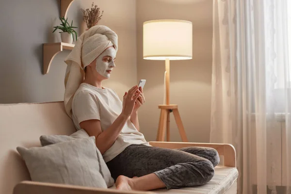 迷人的年轻白种人女人裹着毛巾和面部黏土面具 一边用手机一边在客厅的沙发上放松 一边做美容手术 — 图库照片