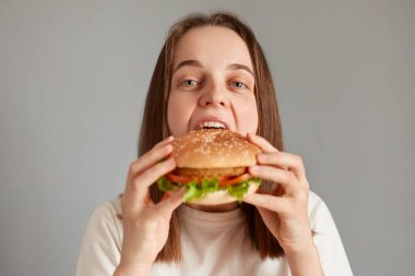 Kahverengi saçlı, çizburgeri zevkle yiyen, hamburger yiyen, fast food restoranından yemek siparişi veren, leziz lezzetlerin tadını çıkaran aç bir kadının kapalı mekândaki fotoğrafı..