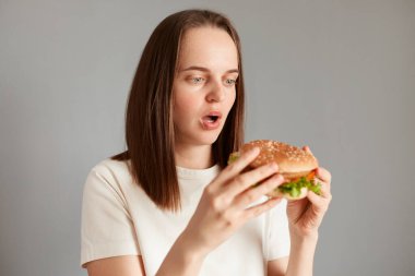 Düzgün beslenme, sağlıklı fast food, sağlıksız seçim. Gri arkaplanda beyaz tişört giyen, el burgerini tutan hayrete düşmüş genç bir kadın..