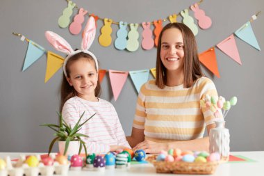 Kapalı alanda gülümseyen çekici anne ve tatlı kızının renkli Paskalya yumurtalarının arasında oturduğu mutlu bir aile Paskalya için hazırlanıyor, tavşan kulağı takan küçük bir kız çocuğu..