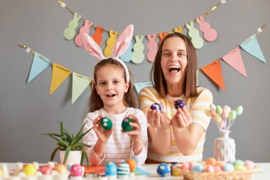 Kahverengi saçlı mutlu anne portresi ve Paskalya yumurtalarını gösteren tavşan kulaklı kızı, Paskalya için hazırlanan tatmin olmuş aile, dekoratif gri duvara poz veriyor..
